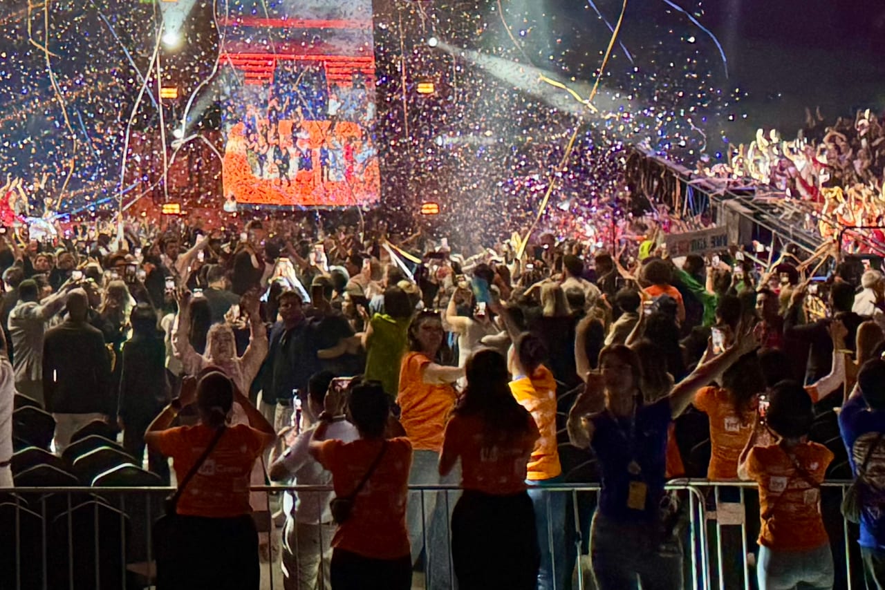 Crew in orange inCruises shirts and attendees with raised arms and phones fill the floor of Belgrade Arena as confetti falls beside a side LED screen under truss lighting