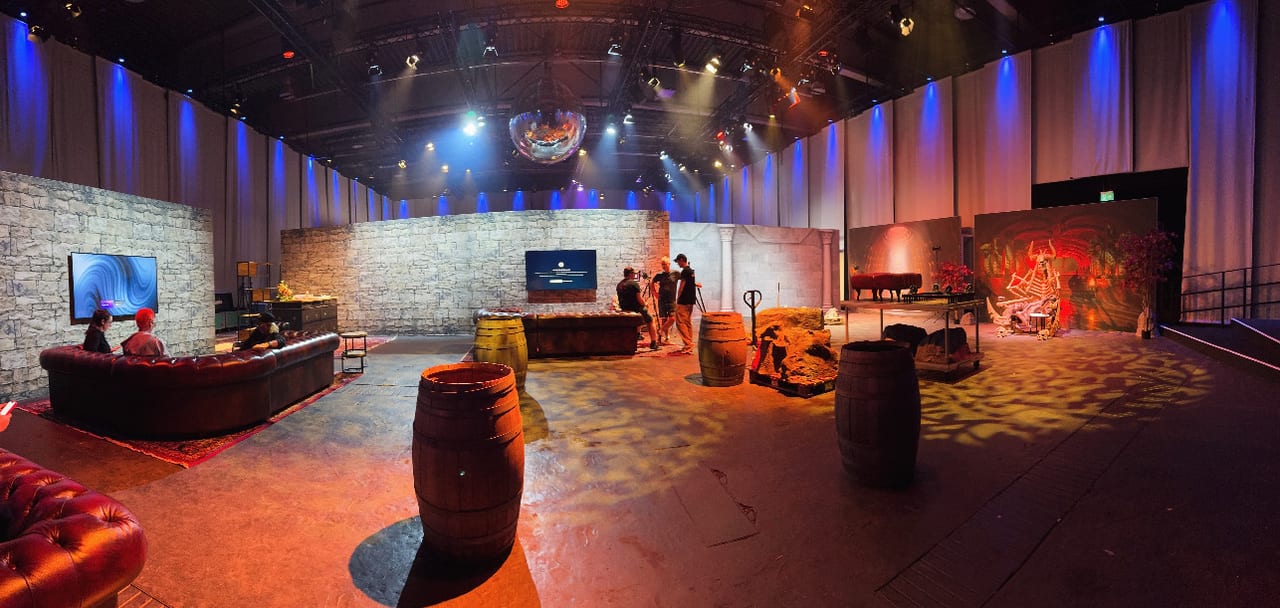 Panorama of the Masters of the Universe lounge set before doors: chesterfield sofas, barrels and stone-wall sets under blue-washed drapes, a disco ball and gobo light patterns across the floor