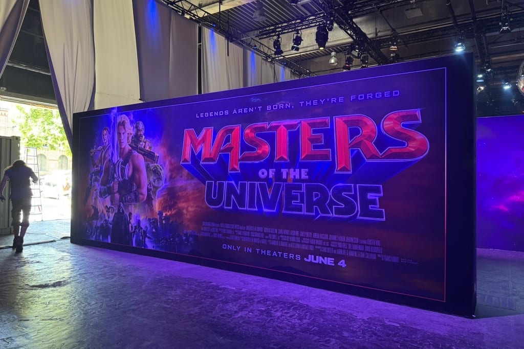A giant Masters of the Universe poster wall, reading "Only in theaters June 4", standing under the lighting rig in the Berlin venue while a crew member walks past