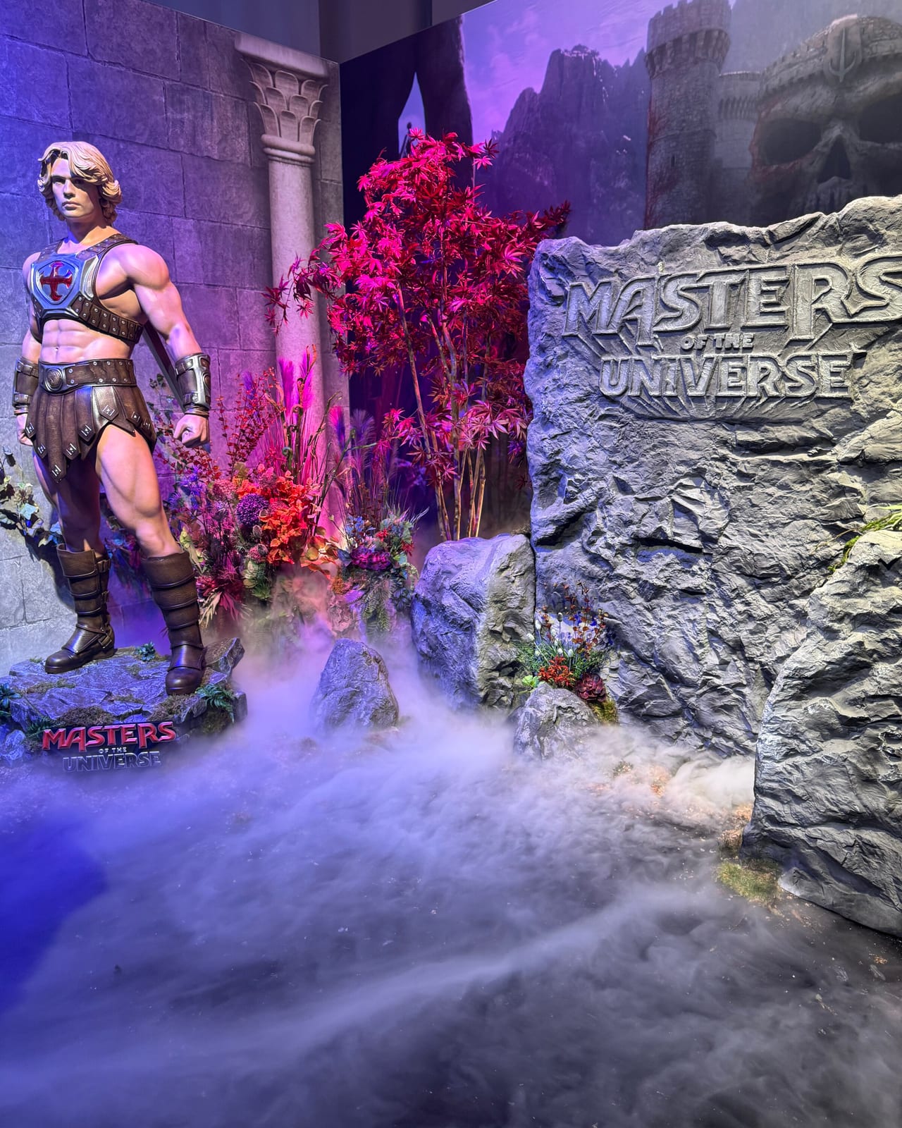 Masters of the Universe creator activation in Berlin: a stone-block castle wall with columns and a lit doorway portal showing a creator posing with a sword, flanked by boulders and magenta foliage