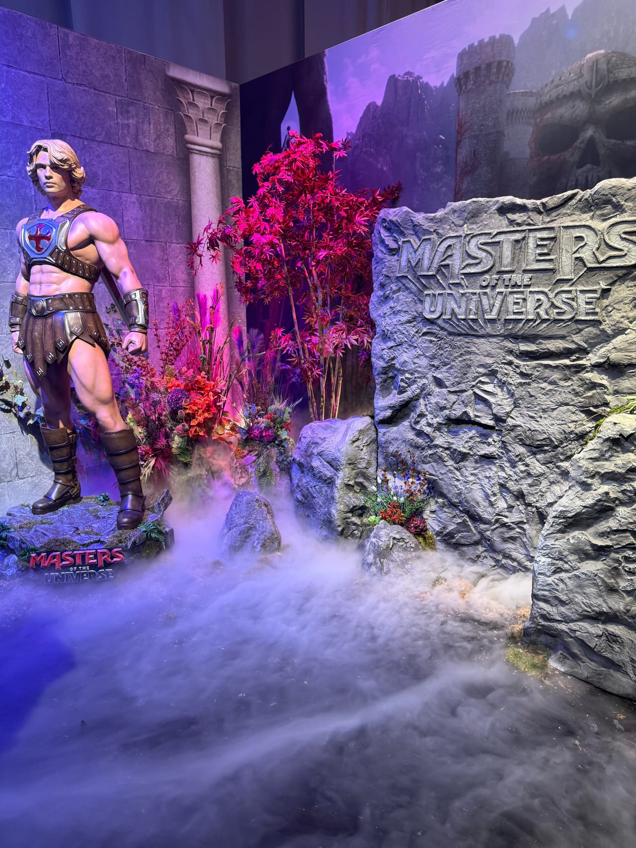 A He-Man statue standing in fog beside the carved Masters of the Universe logo rock, with a red maple and a Castle Grayskull backdrop
