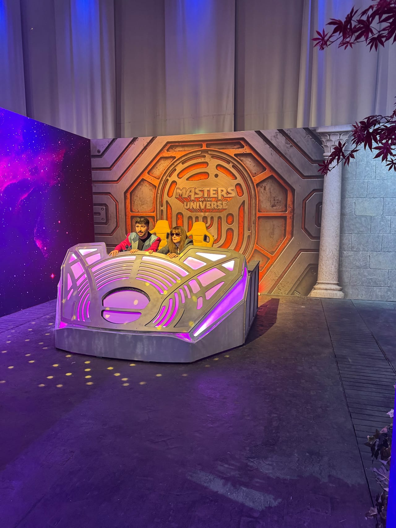 Two creators seated in a glowing purple ship-cockpit photo set in front of the Masters of the Universe emblem