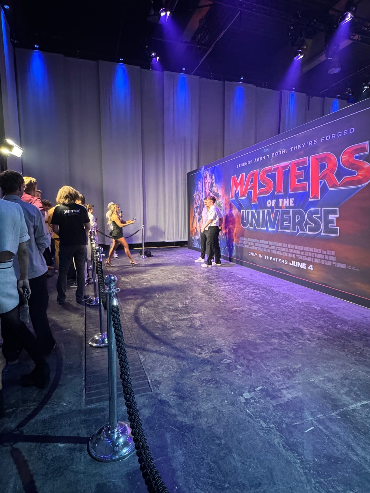Creators queuing behind velvet ropes beside the Masters of the Universe poster wall while others film content in front of it
