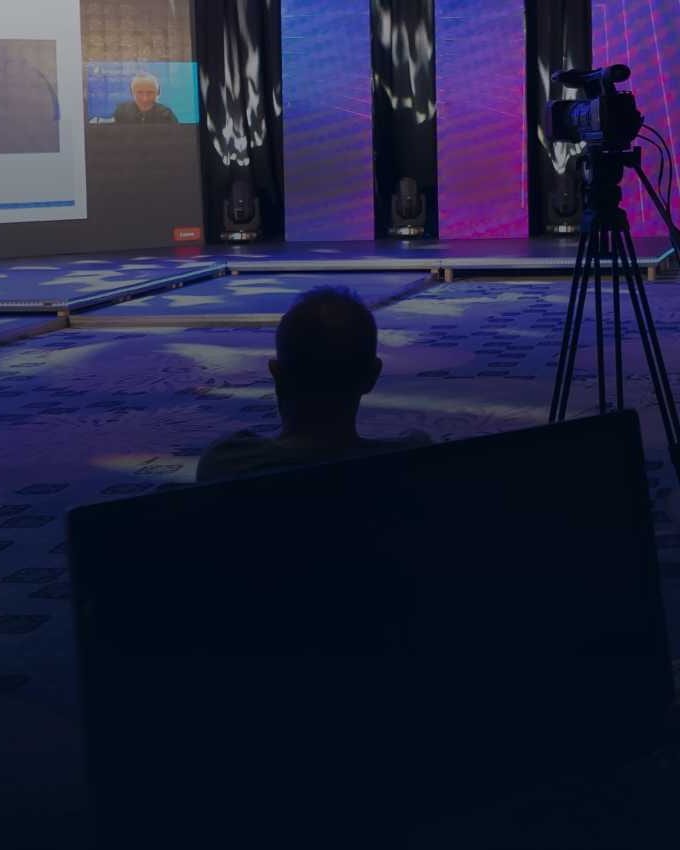 Dark, wide view of a corporate stage with magenta and blue LED walls and a presentation slide, camera operators on tripods and a control desk with monitors in the foreground