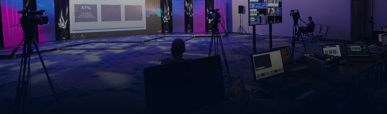 Dark, wide view of a corporate stage with magenta and blue LED walls and a presentation slide, camera operators on tripods and a control desk with monitors in the foreground