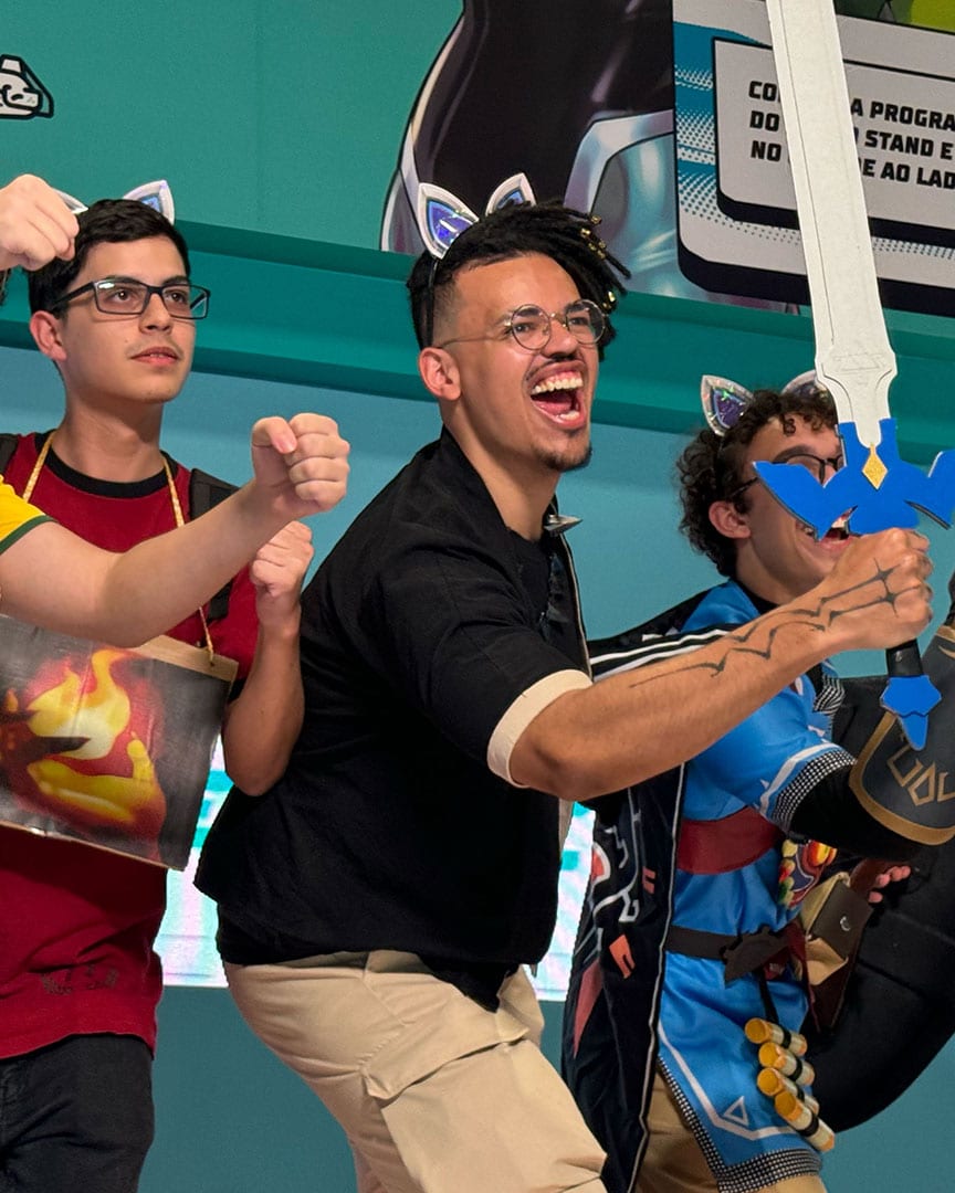 Four fans in a teal-walled venue cheer and pose for a photo, one holding a foam sword and another wearing cat-ear headbands, with a screen and QR code sign behind them