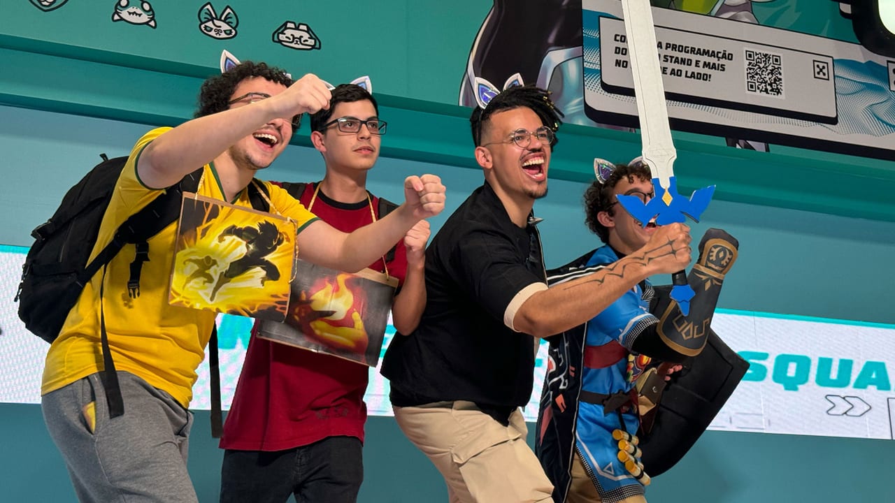Four fans in a teal-walled venue cheer and pose for a photo, one holding a foam sword and another wearing cat-ear headbands, with a screen and QR code sign behind them