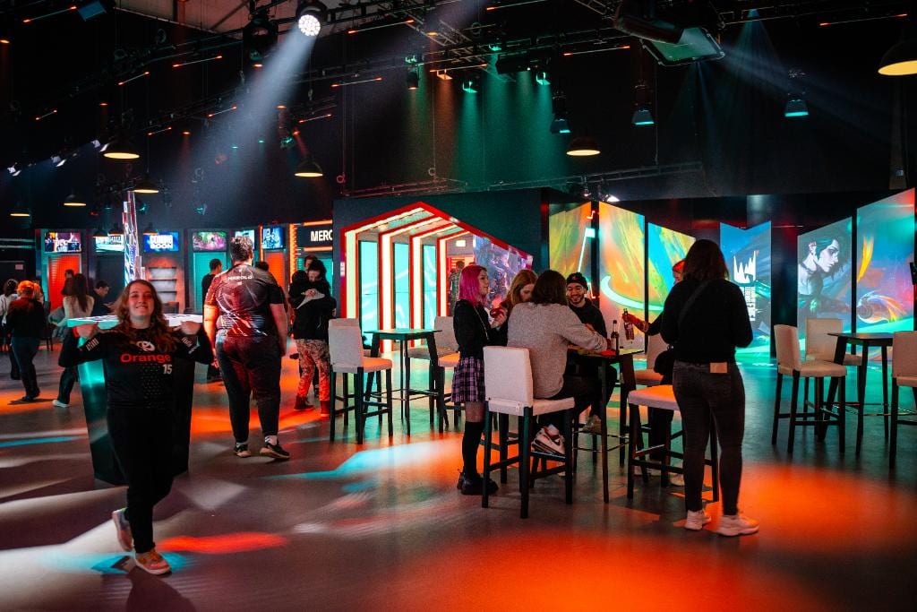 Guests mingle around high tables in a dark hall lit by orange and teal floor light, with glowing portal-style entrances and illustrated character panels along the back wall